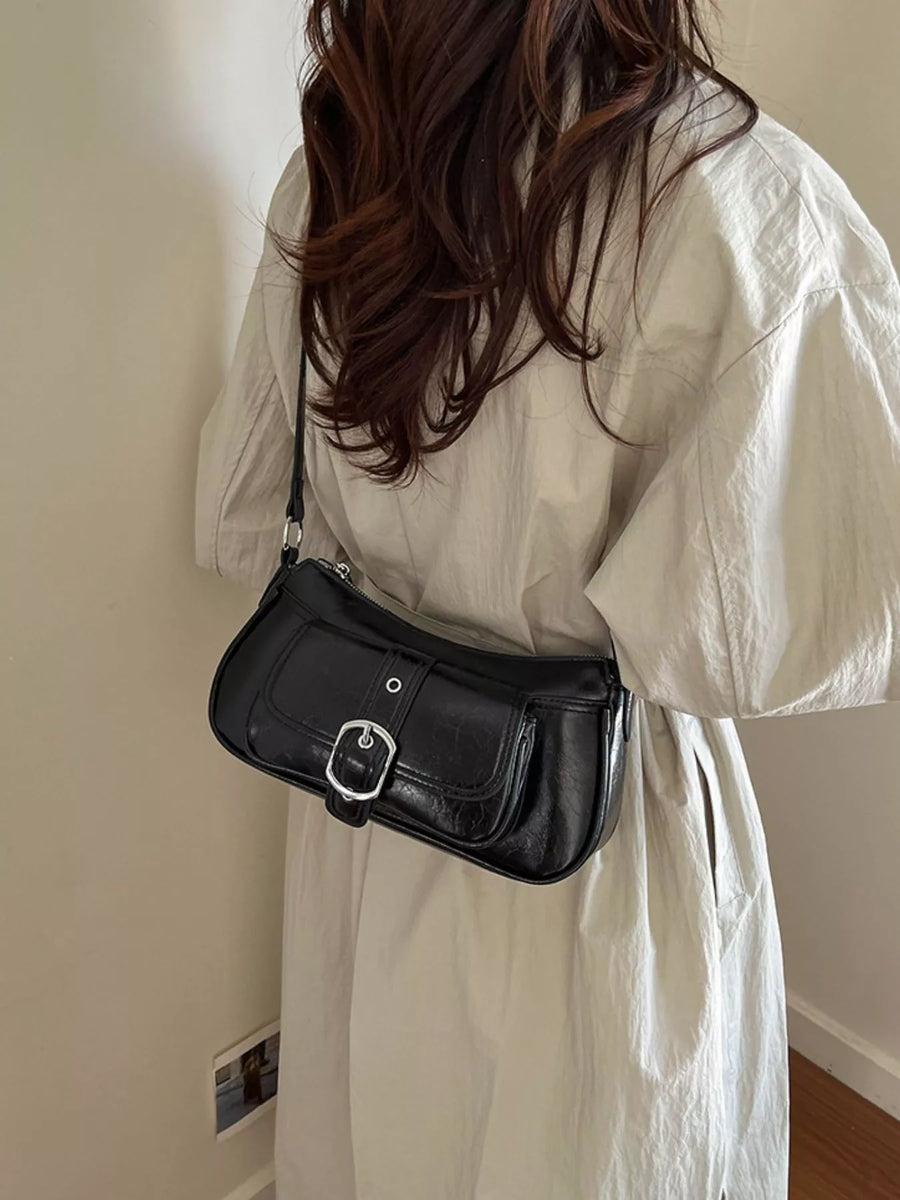 A person wearing a white dress carries a black faux-leather shoulder bag with a silver buckle, shown from behind in an indoor setting.