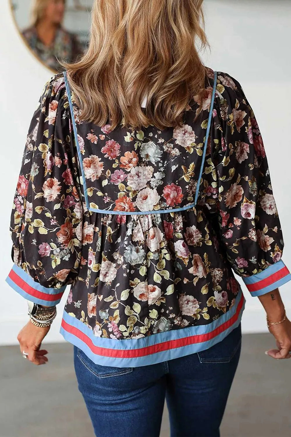 A woman viewed from behind wearing a black floral blouse with puff sleeves and contrasting light blue and red trim, paired with blue jeans.