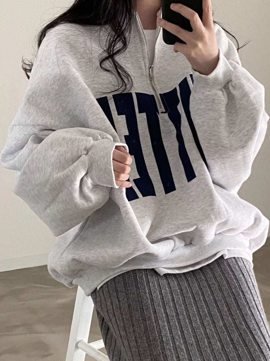 A person wearing an oversized light gray quarter-zip sweatshirt with large navy lettering on the front, paired with a ribbed gray skirt, photographed indoors in a mirror selfie style.
