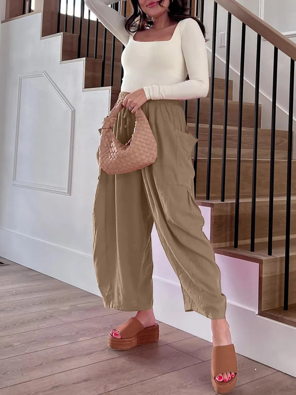 Woman holding a woven handbag in a home setting with wooden stairs.