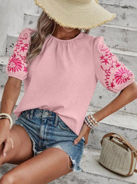 Person wearing a pink top with floral embroidery and denim shorts, sitting on a wooden floor.