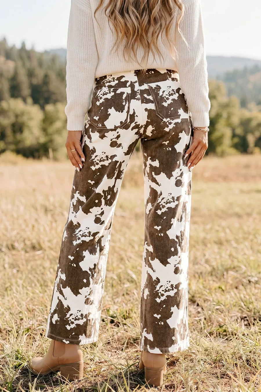 A person standing outdoors in a grassy field wearing brown and white tie-dye wide-leg pants, a white ribbed sweater, and tan ankle boots.