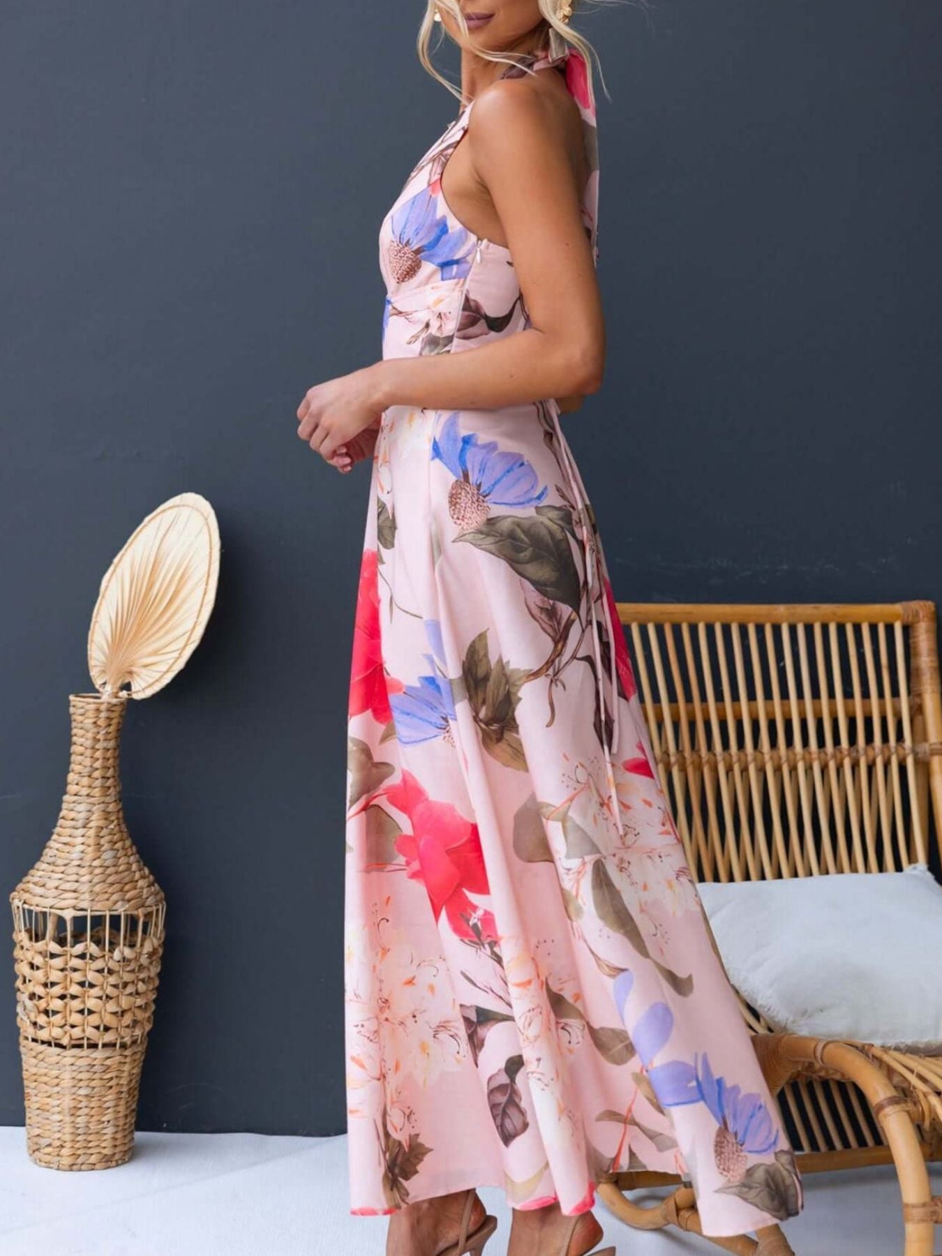 Woman wearing a floral dress standing next to a wicker chair and decorative vase against a dark wall.