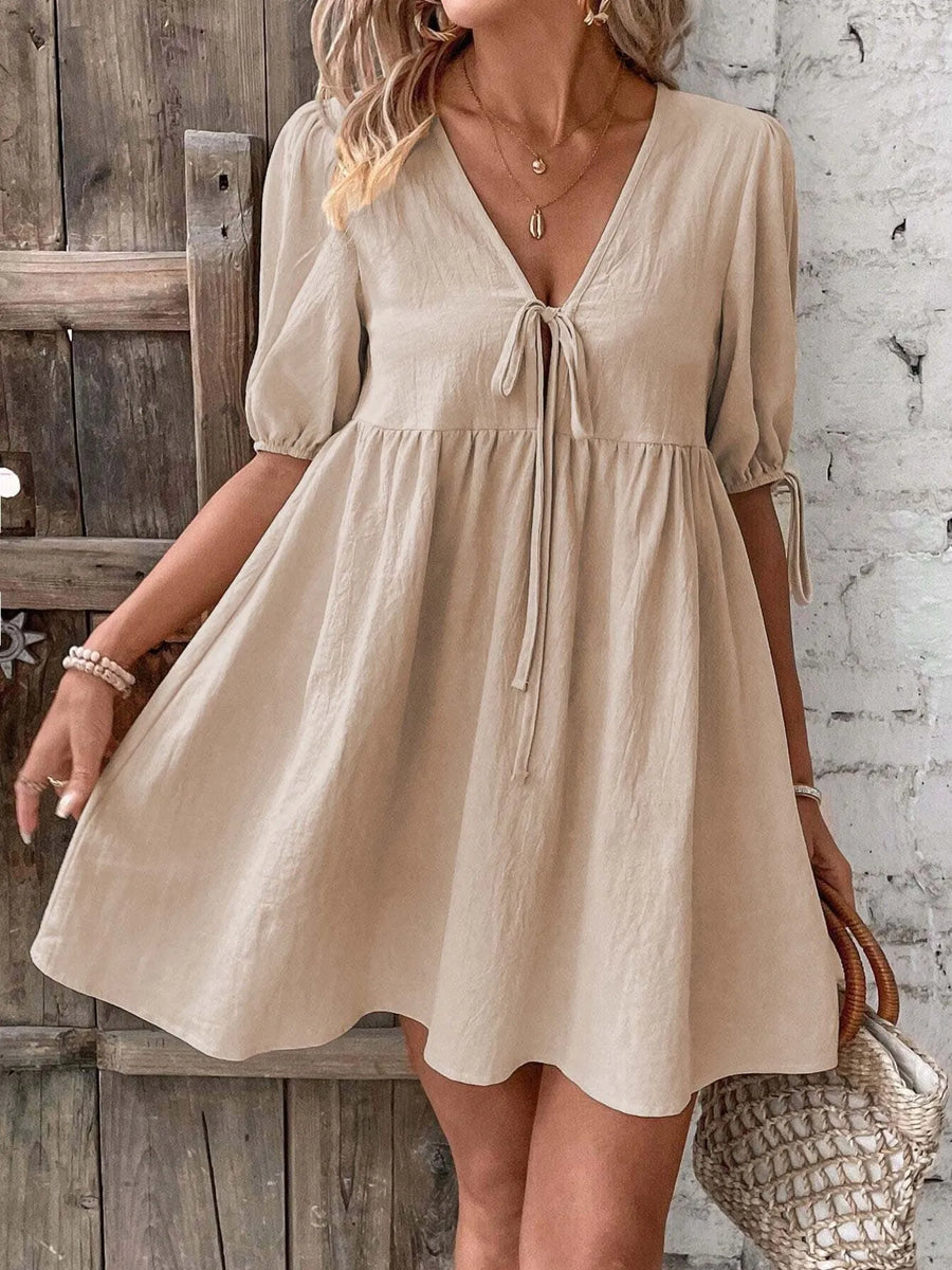 Woman wearing a beige dress against a rustic wooden and stone wall.