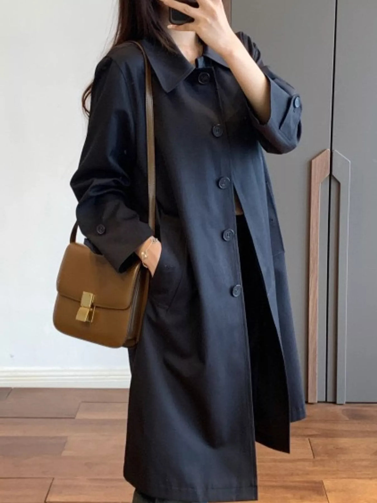 A woman wearing a long black trench coat and carrying a brown leather shoulder bag with a gold clasp, standing in an indoor setting with a wooden floor.