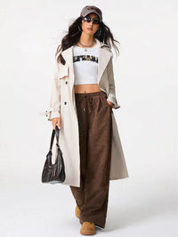 Woman wearing a white trench coat, white crop top, brown wide-leg pants, and black sunglasses on a white background