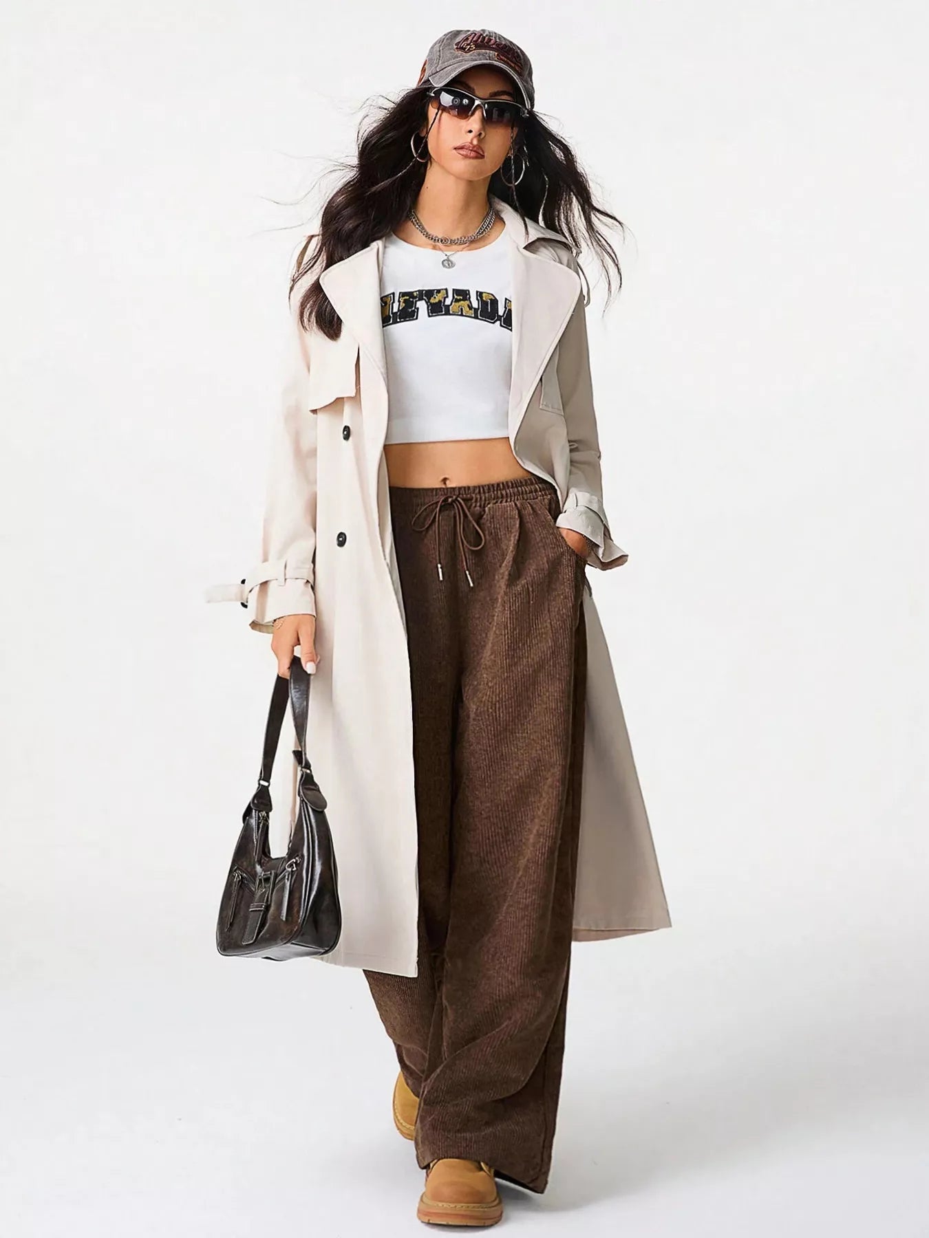 Woman wearing a white trench coat, white crop top, brown wide-leg pants, and black sunglasses on a white background