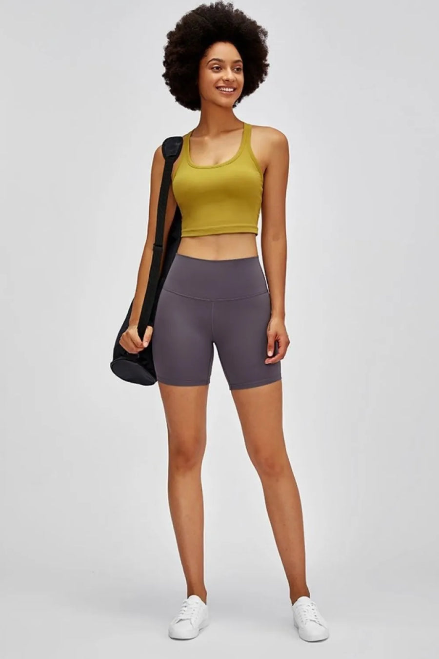 Woman wearing a yellow tank top and purple shorts with a black bag on a white background