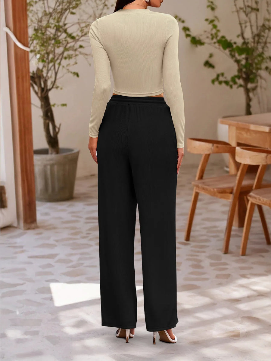 Person wearing a beige top and black pants in an indoor setting with wooden chairs and plants.