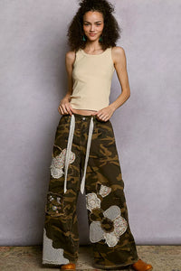 Woman wearing a beige tank top and camouflage pants with lace details against a gray background