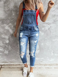 Person wearing a red top and blue ripped overalls against a gray concrete wall.