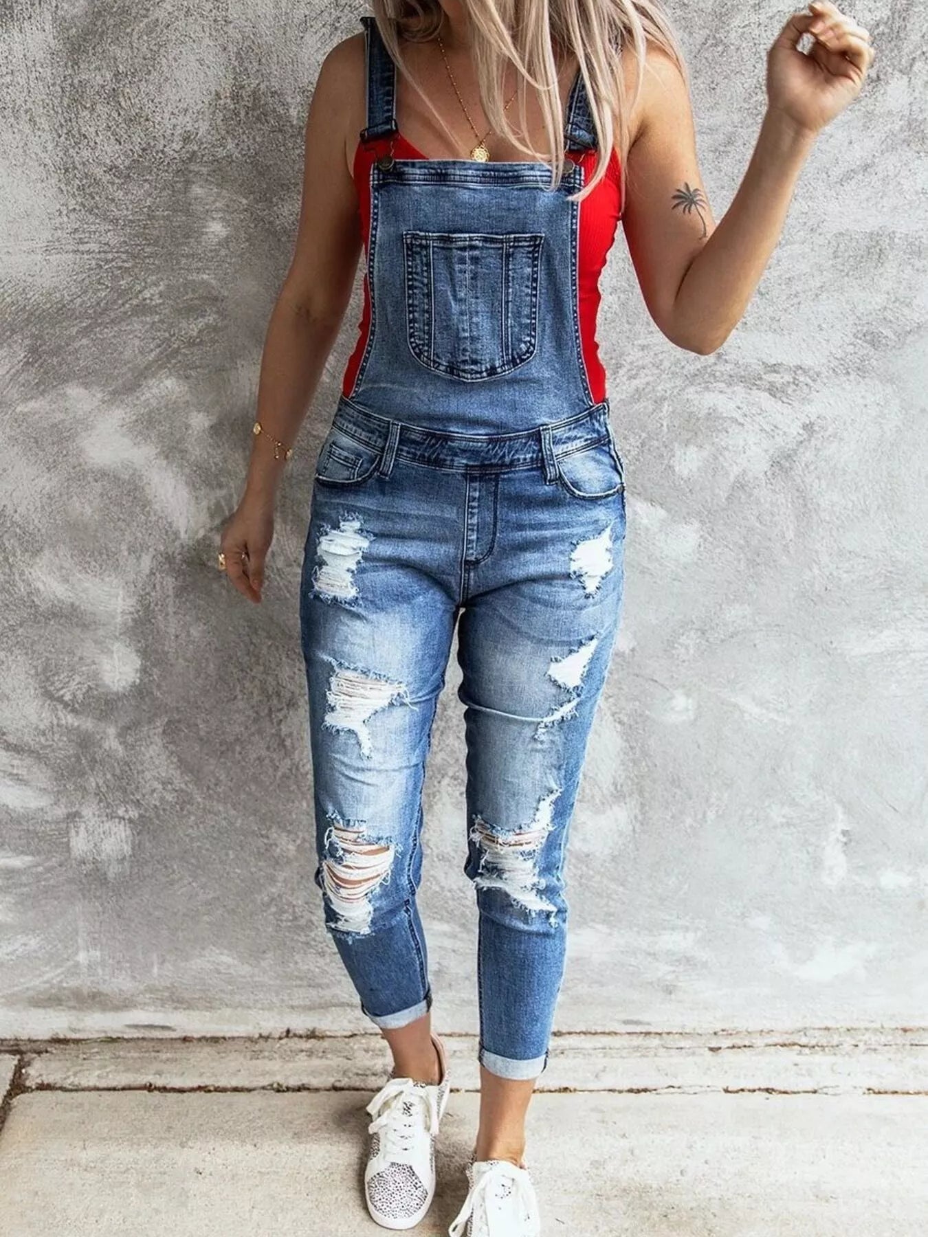 Person wearing a red top and blue ripped overalls against a gray concrete wall.