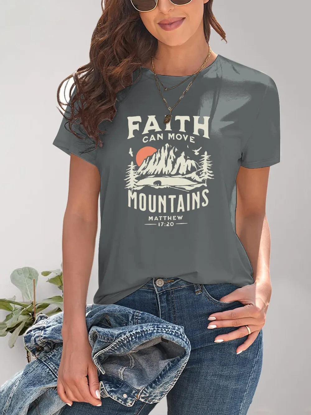Person wearing a gray t-shirt with 'Faith Can Move Mountains' design, holding jeans.