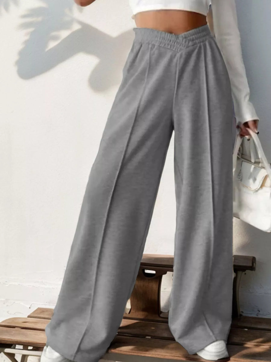 Person wearing gray wide-leg pants and a white top, sitting on a wooden bench.