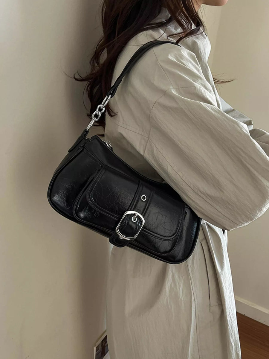 A woman wearing a light beige trench coat carries a black faux-leather shoulder bag with a silver buckle and chain strap against a plain beige wall.