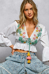 Woman wearing a white blouse with colorful embroidery and blue jeans against a gray background