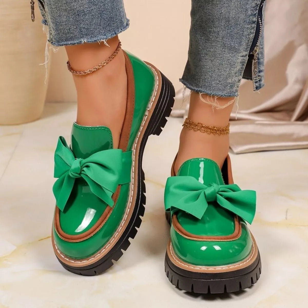 A pair of bright green patent leather loafer-style shoes with large satin bows and chunky black lug soles, shown on a model's feet with frayed blue jeans.