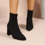 Black ankle boots with a pointed toe and block heel, shown on a mannequin leg against a neutral beige background.