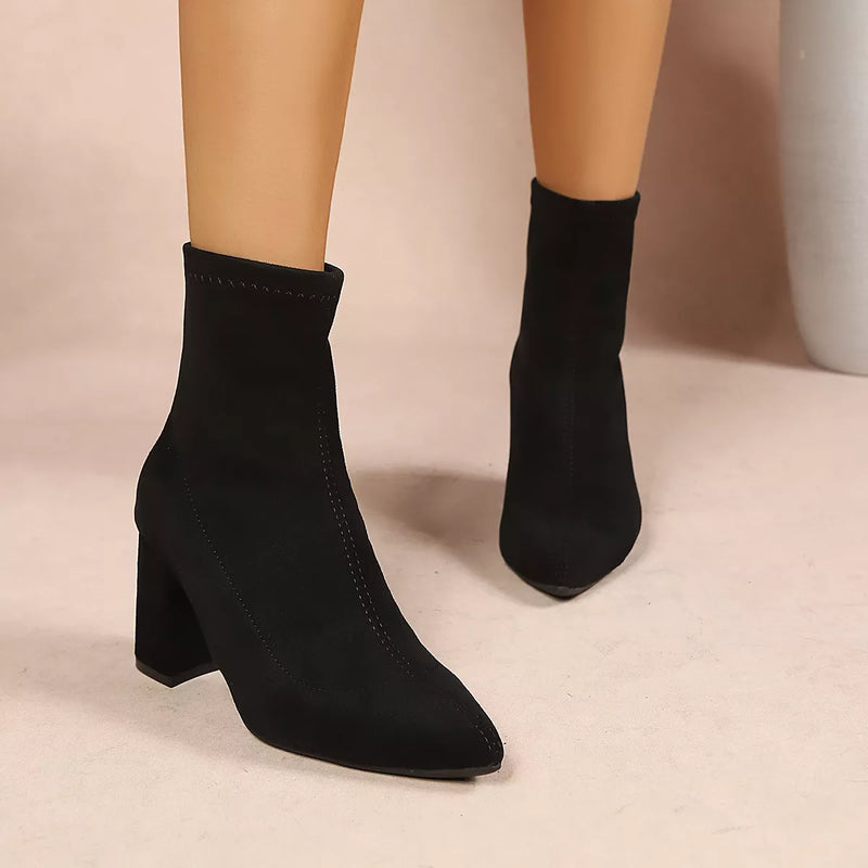 Black ankle boots with a pointed toe and block heel, shown on a mannequin leg against a neutral beige background.