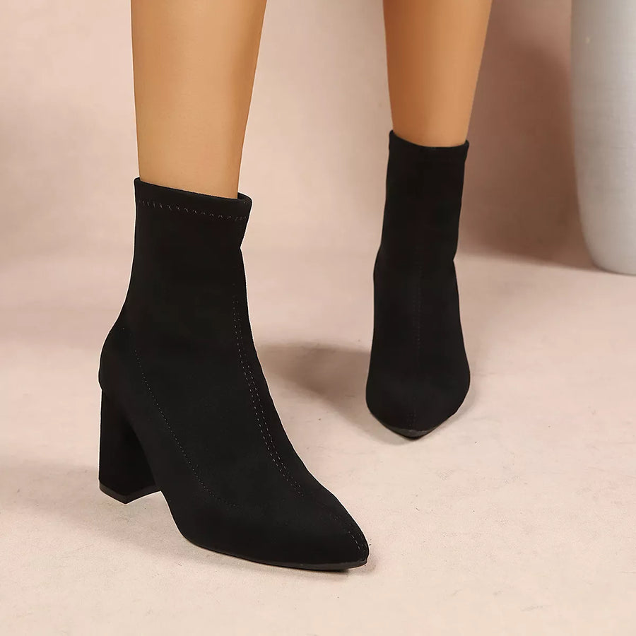 Black ankle boots with a pointed toe and block heel, shown on a mannequin leg against a neutral beige background.