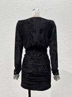 A black long-sleeve mini dress with floral lace embroidery and ruching, shown from the back on a mannequin against a textured white wall.