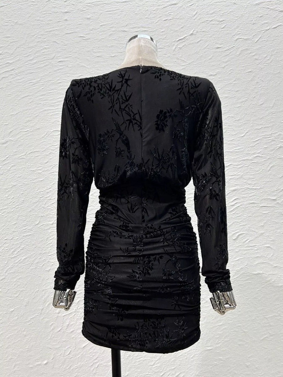 A black long-sleeve mini dress with floral lace embroidery and ruching, shown from the back on a mannequin against a textured white wall.