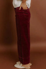 A person wearing dark burgundy velvet wide-leg pants, styled with a white top and white flat shoes, standing against a warm brown studio backdrop.