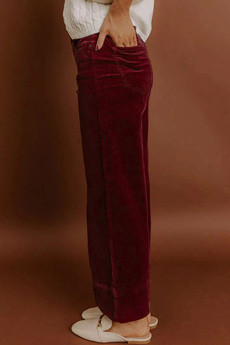 A person wearing dark burgundy velvet wide-leg pants, styled with a white top and white flat shoes, standing against a warm brown studio backdrop.