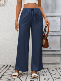 Person wearing navy wide-leg pants with a white top and brown sandals, standing against a gray wooden panel background.