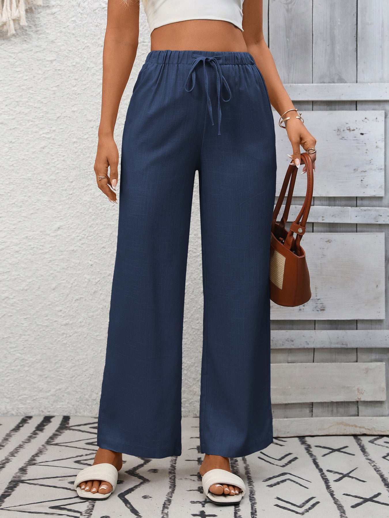 Person wearing navy wide-leg pants with a white top and brown sandals, standing against a gray wooden panel background.