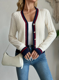 Woman wearing a white cable knit cardigan with blue and red accents, holding a beige handbag.
