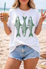 Person on a beach wearing a white t-shirt with green fish graphics and denim shorts.