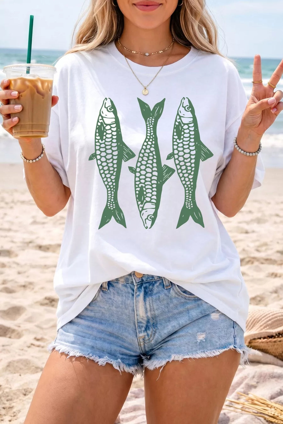 Person on a beach wearing a white t-shirt with green fish graphics and denim shorts.