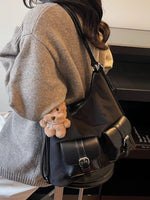 Person wearing a gray sweater with a black leather bag and a small teddy bear attached, sitting on a bed.