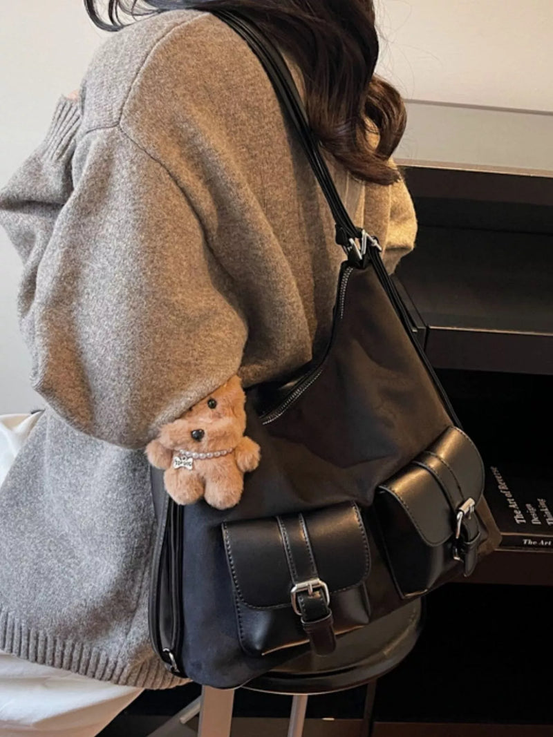 Person wearing a gray sweater with a black leather bag and a small teddy bear attached, sitting on a bed.