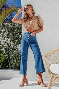 Woman wearing a beige top and blue jeans standing outdoors with plants and a chair in the background.