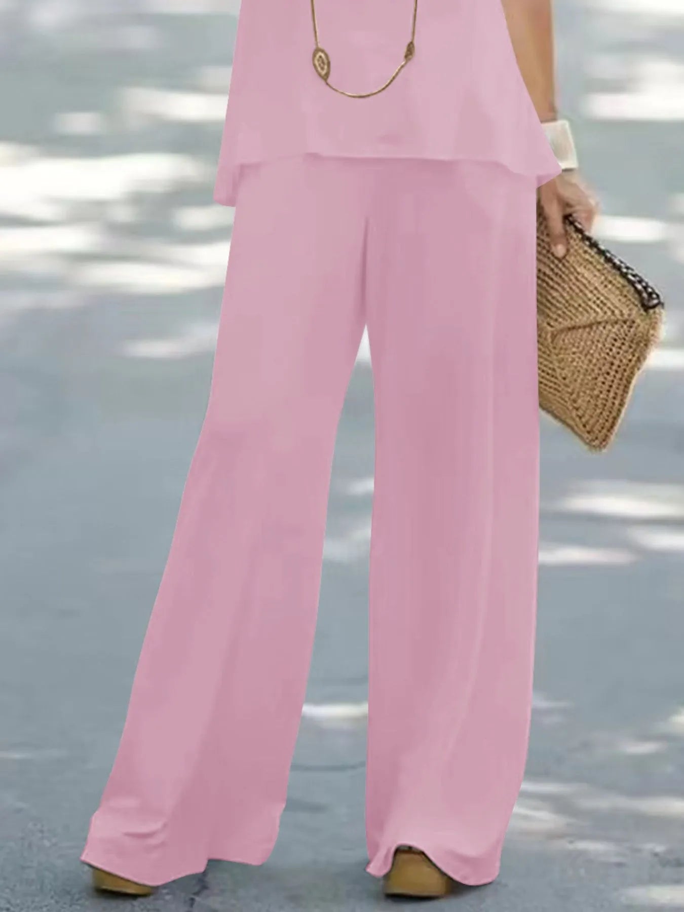 Person wearing a pink outfit with wide-leg pants and holding a straw bag on a light gray background.