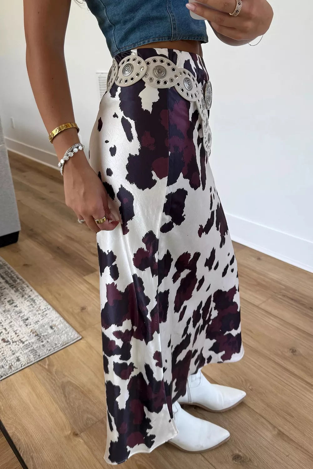 A person wearing a long white skirt with a dark purple and black cow-print pattern, styled with a denim top, white boots, and a decorative belt in an indoor room setting.