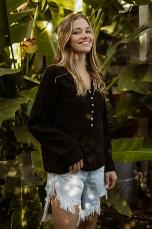 Woman standing among large green leaves wearing a black jacket and denim shorts.
