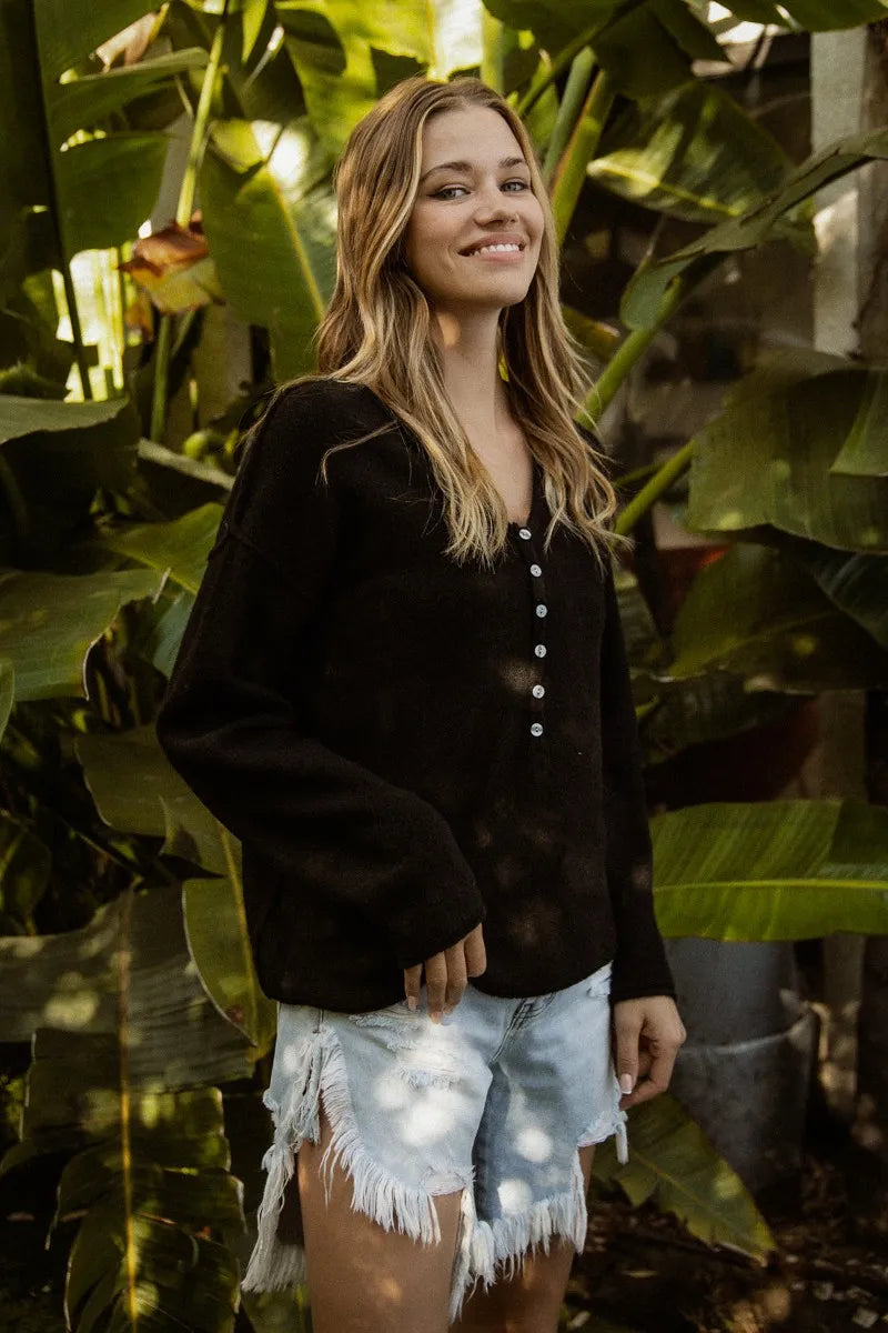 Woman standing among large green leaves wearing a black jacket and denim shorts.