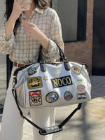 Person holding a silver bag with multiple patches, including 'NOCO', against a neutral background.