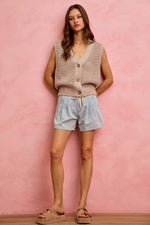 Woman wearing a beige knitted vest and light blue shorts against a pink background