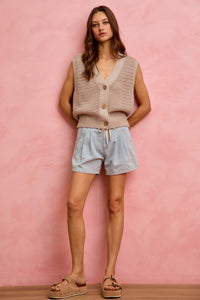 Woman wearing a beige knitted vest and light blue shorts against a pink background