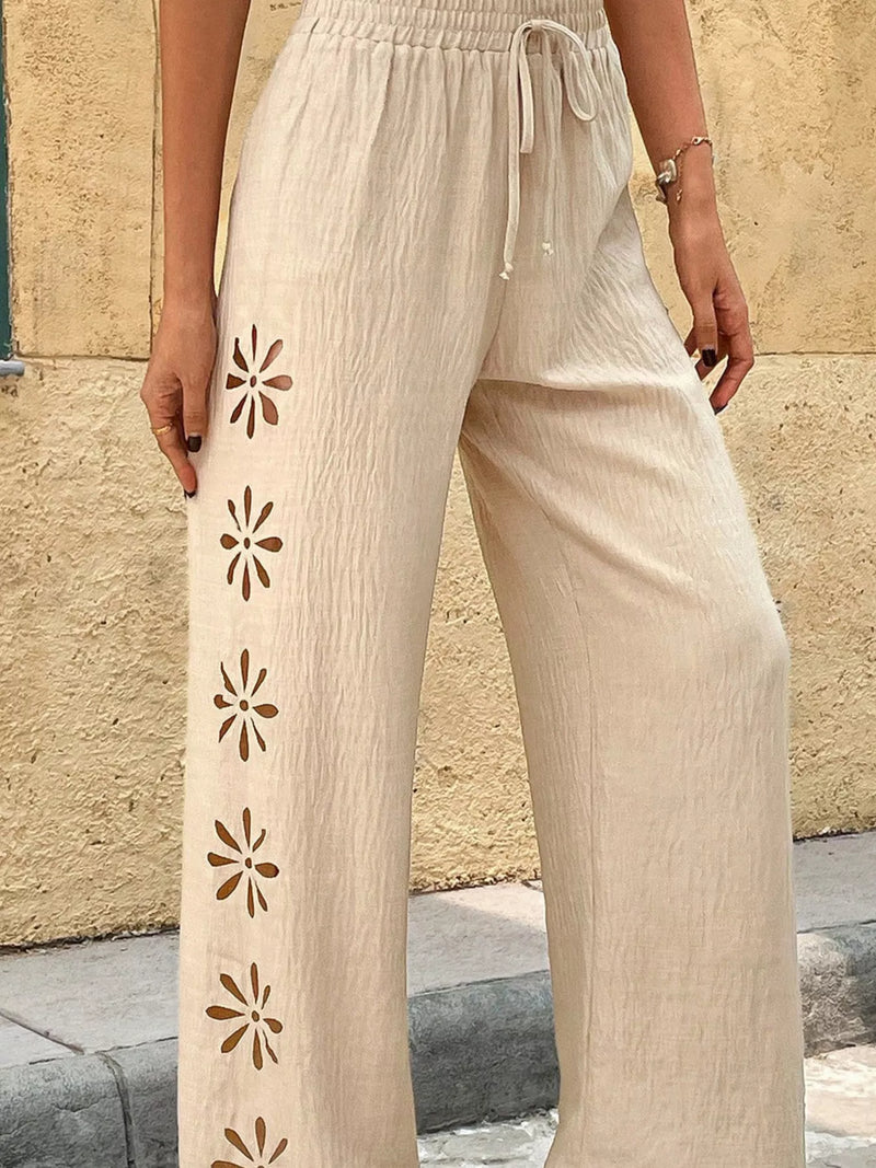Beige pants with floral cut-out design worn by a person against a stone wall.