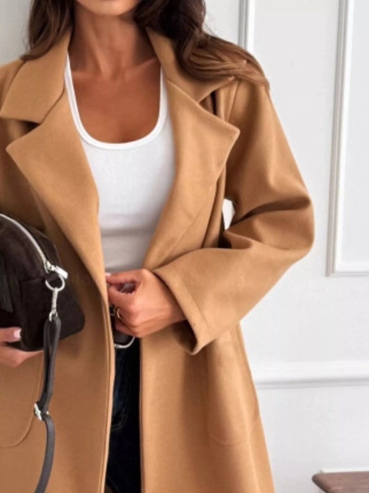 Person wearing a tan coat over a white top, holding a black handbag indoors.