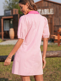 Woman wearing a pink and white striped dress standing outdoors with a wooden building in the background.