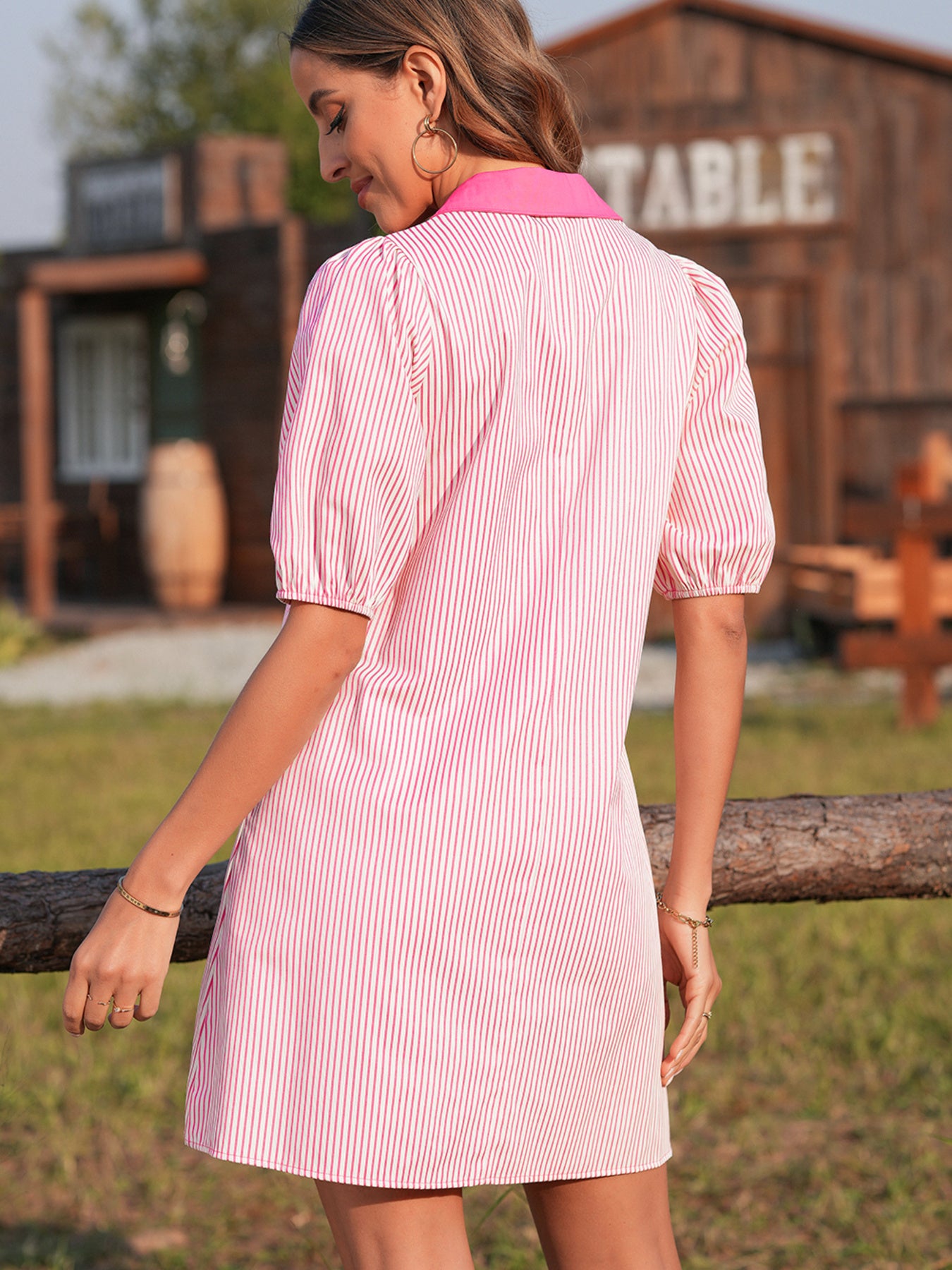 Woman wearing a pink and white striped dress standing outdoors with a wooden building in the background.