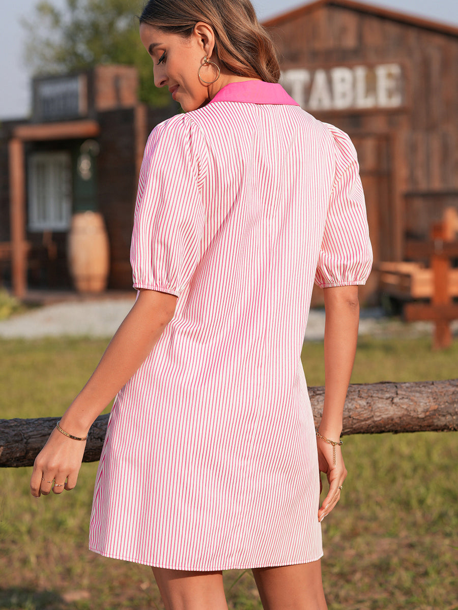 Woman wearing a pink and white striped dress standing outdoors with a wooden building in the background.