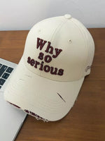 Beige cap with 'Why so serious' text on a wooden surface next to a laptop.