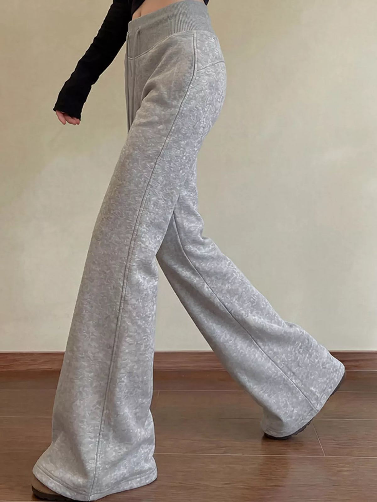 A person wearing light heather gray wide-leg sweatpants, shown from the waist down in a side profile against a beige wall and wooden floor.
